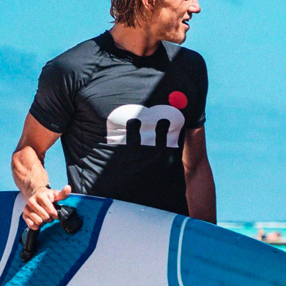A man with a surfboard wearing a Mistral shirt